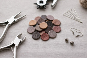 Flat lay of leather discs in brown shades with jewelry tools, earring hooks, pins, and metal beads on a neutral fabric background