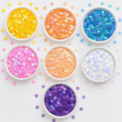Seven white ceramic bowls filled with 7mm flat daisy flower sequins in yellow AB, orange AB, blue AB, pink AB, peach AB, white AB and purple AB iridescent colours arranged in a heart shape on white background
